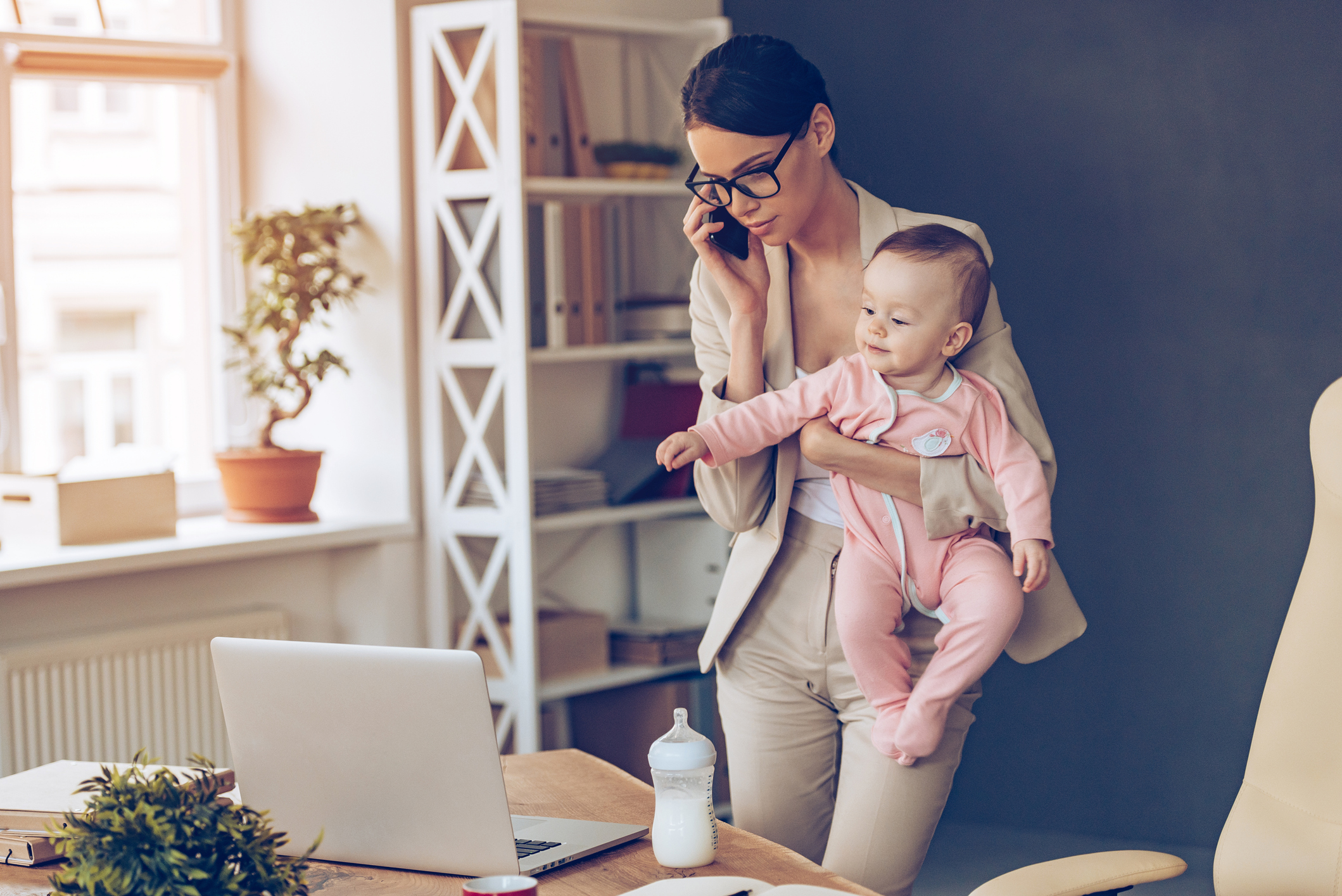 A working mother talking on a cell phone while looking at a laptop and holding a baby.