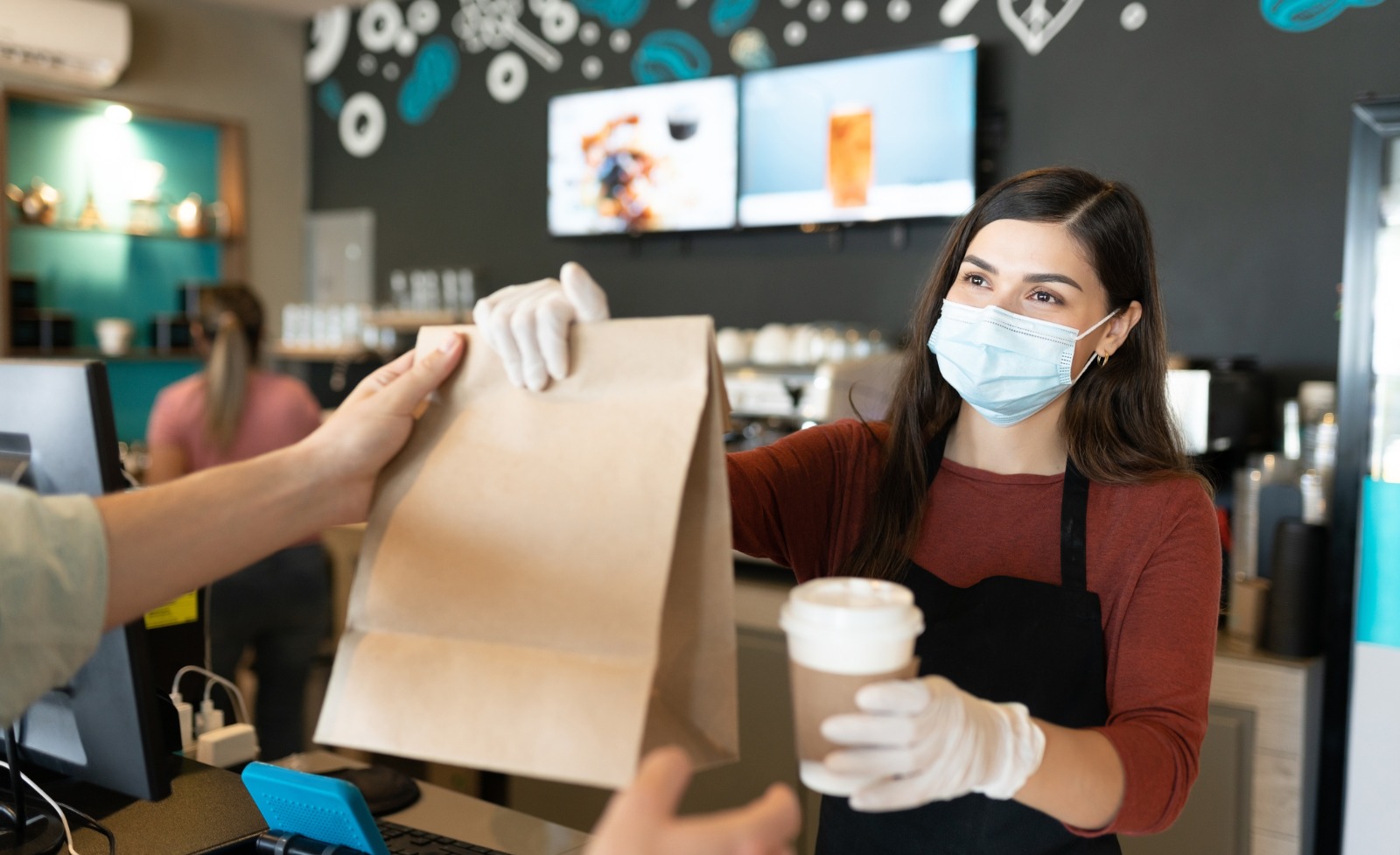 A cashier wearing a mask passing a paper bag and cup of coffee to a customer.