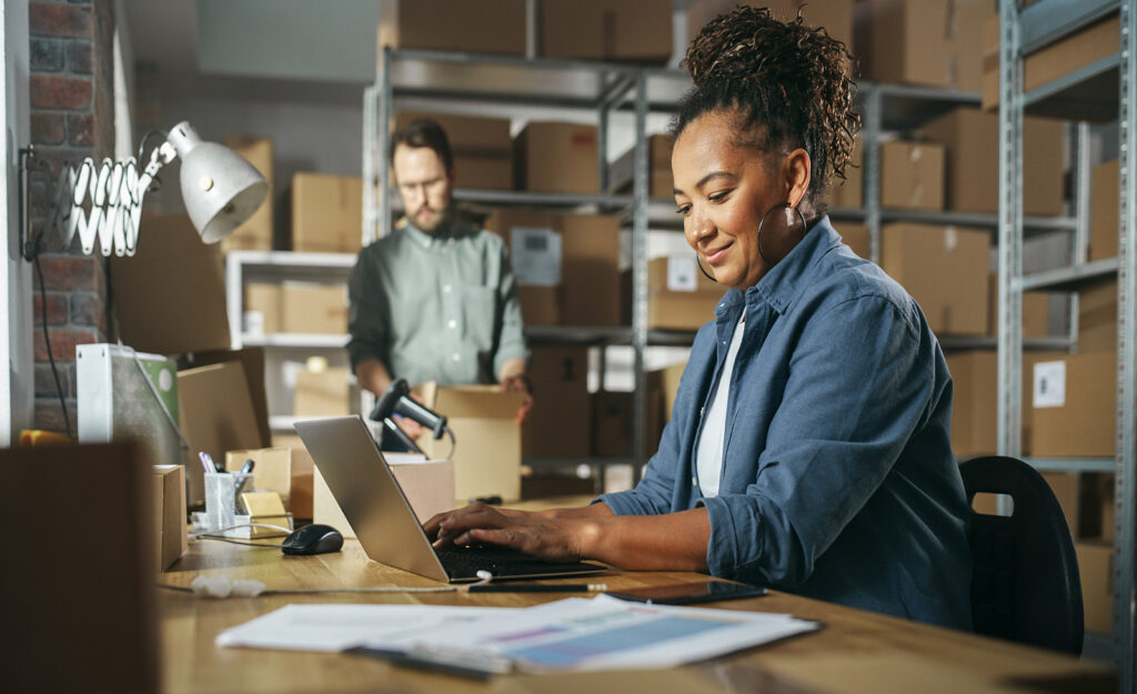 An African American woman typing on a laptop in a warehouse full of boxes on shelves and an employee holding a box in the background.