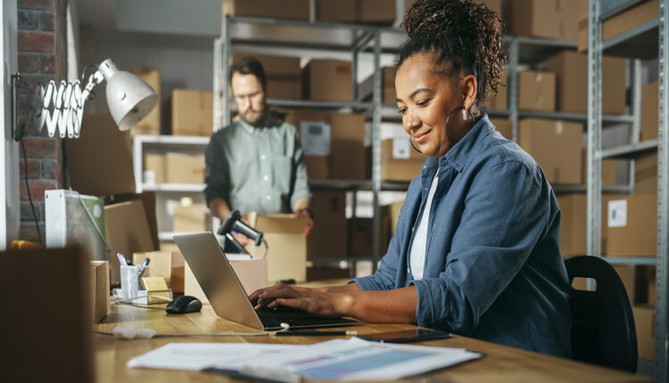 An African American woman typing on a laptop in a warehouse full of boxes on shelves and an employee holding a box in the background.