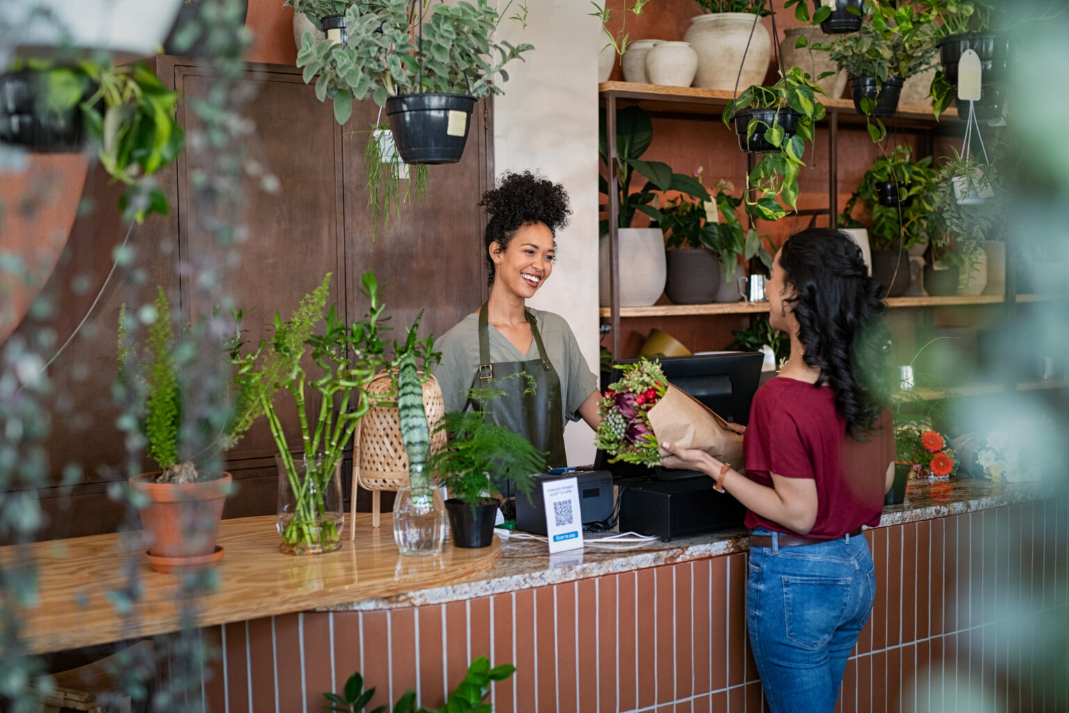 A smiling woman is selling a bouquet to a customer while standing in a flower shop.